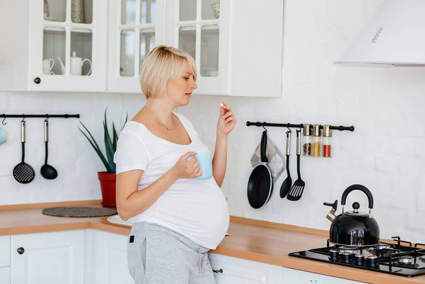 Een jong blond zwanger meisje in een wit T-shirt en grijze broek staat in de keuken met een thermometer en pillen in haar handen - Foto, afbeelding