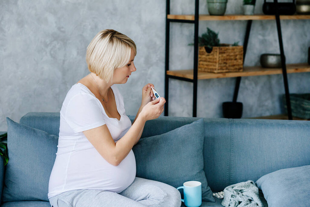 Молодая блондинка беременная сидит на сером диване в белой футболке и серых брюках и измеряет температуру с целью выявления заболевания
 - Фото, изображение