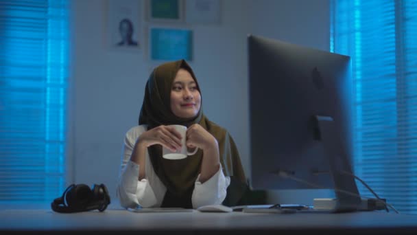 Sleepy Asian Muslims wearing dark headscarves drinking coffee when working from home in a modern interior design workspace with the warm blue light from the windows - Felvétel, videó