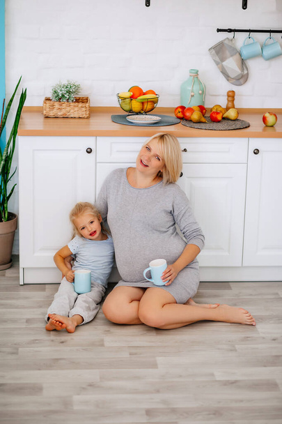 Μια έγκυος κοπέλα κάθεται με μια μικρή ξανθιά κοπέλα στο πάτωμα της κουζίνας με κύπελλα στα χέρια της - Φωτογραφία, εικόνα