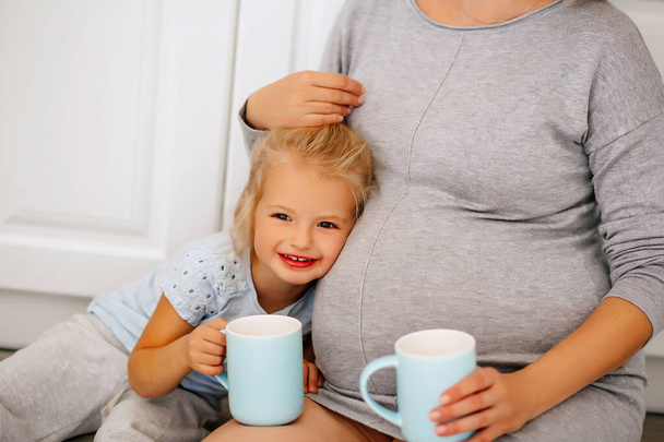 Een zwanger meisje zit met een klein blond meisje op de vloer in de keuken met kopjes in haar handen - Foto, afbeelding