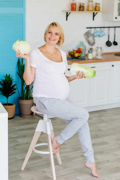 Pregnant blonde in the kitchen at the table parses green cabbage - Foto, afbeelding