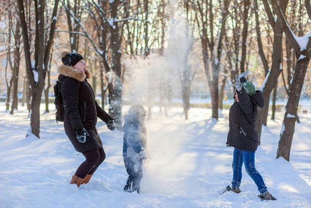 Mutter mit zwei Söhnen spielt im Schnee im Park. Gefriertag. Schnee schmeißen. Im Hintergrund stehen Bäume. Ruhetag. Freizeit. Familienzeit. Wintersonne. - Foto, Bild