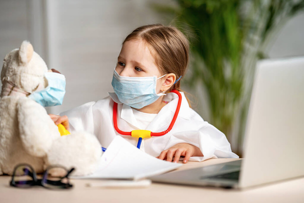 charming little girl in a white coat and mask, using a stethoscope, listens to Teddy bear. Cute little preschooler in uniform playing game of doctor patient in clinic, concept of children's health - Photo, Image