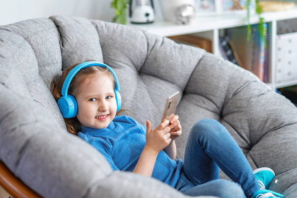 little girl with headphones and holding smartphone in her hands lies on large round armchair and looks at screen frame and smiles listens to music. Side view leisure Concept during covid-19 quarantine - Foto, afbeelding