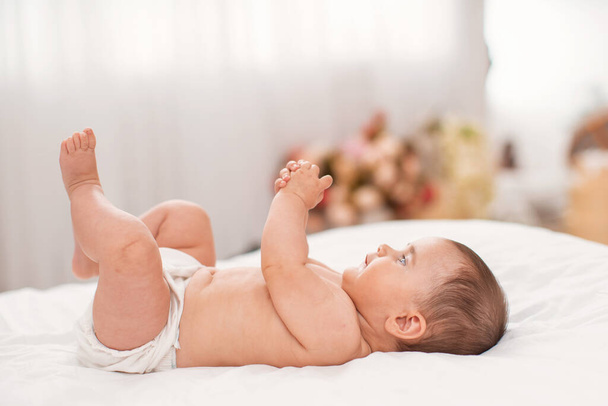 Cute 4 month old baby is lying on his back on the bed and playing with his hands. Bed with light linen. The room is cozy, light and lots of flowers in vases. - Fotó, kép