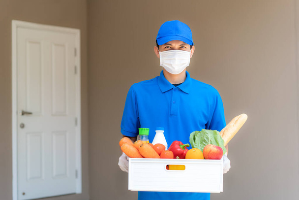 Asian delivery man wearing face mask and glove with groceries box of food, fruit, vegetable and drink standing in front of the customer house during time of home isolation. - Valokuva, kuva