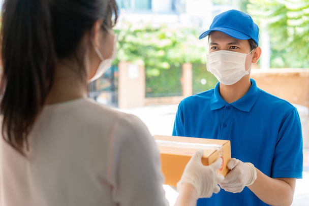 Azjatycki kurier w masce i rękawiczce w niebieskim mundurze trzymający kartonowe pudełka w domu i kobieta przyjmująca dostawę pudełek od kuriera podczas wybuchu epidemii COVID-19. - Zdjęcie, obraz