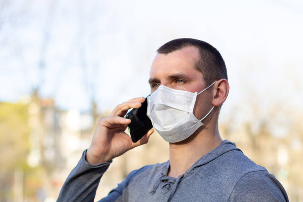 Yüzünde tıbbi maske olan beyaz bir adam telefonla arıyor. Koruyucu maskeli adam dışarıda virüsten korunuyor.. - Fotoğraf, Görsel