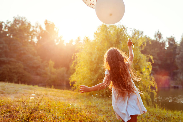 Ευτυχισμένο κοριτσάκι που τρέχει με μπαλόνια στο χέρι. Το παιδί διασκεδάζει στο πάρκο το ηλιοβασίλεμα. Δραστηριότητες σε εξωτερικούς χώρους - Φωτογραφία, εικόνα