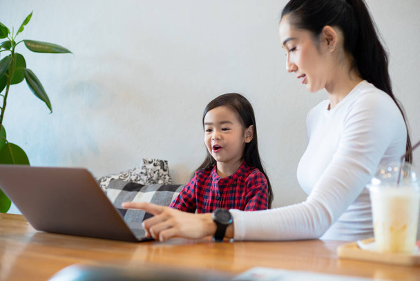 Asian mothers are teaching their daughters to read a book and use notebooks and technology for online learning during school holidays at home. Educational concepts and activities of the family - Photo, Image