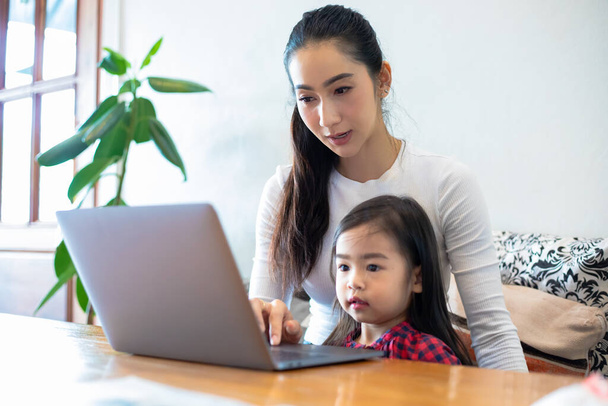 Asian mothers are teaching their daughters to read a book and use notebooks and technology for online learning during school holidays at home. Educational concepts and activities of the family - Foto, Bild