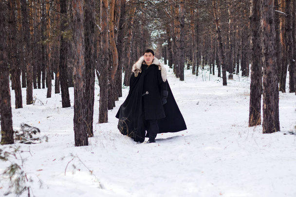 Um guerreiro de roupas pretas acolchoadas em um manto preto com um colarinho de pele branca e uma longa espada em suas mãos caminha ao longo da estrada na floresta
. - Foto, Imagem
