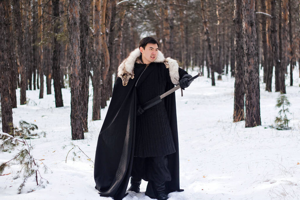 A warrior in black quilted clothes in a black cloak with a white fur collar and a long sword in his hands walks along the road in the forest. - Fotó, kép