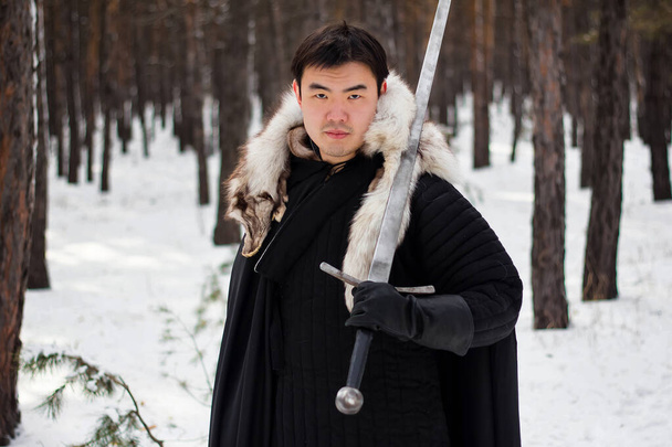 Válečník v černém prošívaném oblečení v černém plášti s bílým kožešinovým límcem a dlouhým mečem v rukou kráčí po cestě v lese. - Fotografie, Obrázek