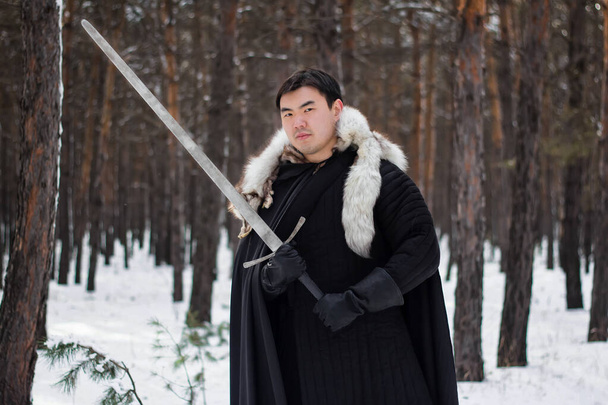A warrior in black quilted clothes in a black cloak with a white fur collar and a long sword in his hands walks along the road in the forest. - Foto, Imagem