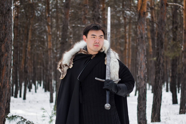 Egy fekete ruhás harcos fekete köpenyben, fehér gallérral és hosszú karddal a kezében sétál az úton az erdőben.. - Fotó, kép