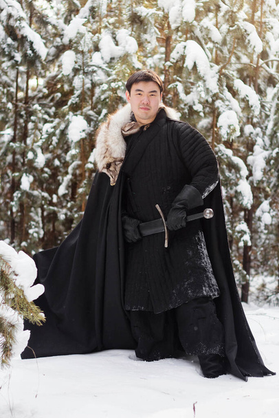A warrior wearing black quilted clothes in a black cloak with a white fur collar and a long sword in his hands is walking along deep snow against the backdrop of a pine forest. - Foto, Imagem