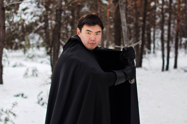 Portrét válečníka v černých prošívaných šatech, kožených rukavicích, plášti s kapucí a mečem v rukou na pozadí zimního lesa. - Fotografie, Obrázek