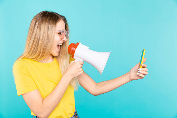 Angry young woman shout with megaphone to mobile phone on blue background - Photo, Image