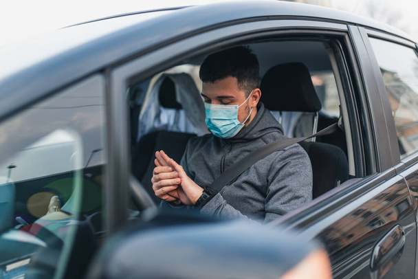 Ein Mann sitzt im Auto und wäscht sich die Hände mit antiseptischem Gel. Gesundheitskonzept im Auto. Die Maske ist weiß im Gesicht. Coronavirus, Krankheit, Infektion, Quarantäne, Covid-19 - Foto, Bild