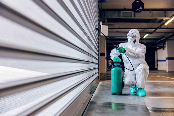Werknemer in steriel beschermingspak en masker hurkend steriliserende deur van een garage van corona virus / covid-19. - Foto, afbeelding
