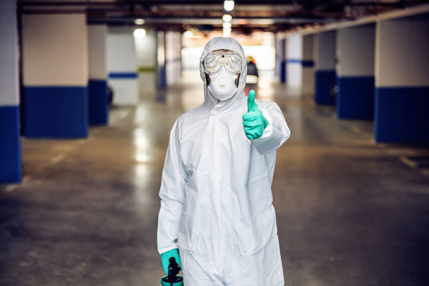 Man in sterile uniform and mask standing in underground garage and showing thumbs up. He sterilized all surface in underground garage. - Photo, Image