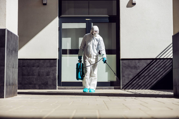 Full length of man in sterile uniform and mask sterilizing surface outdoors from corona virus, fungus and disease. - Fotoğraf, Görsel