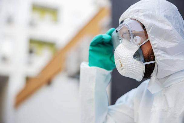Profile of man in protective sterile uniform and mask putting glasses while standing outdoors. - Photo, image