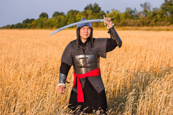 A medieval nomad warrior in oriental armor on the background of a wheat field. Standing in a fencing rack. - Фото, зображення