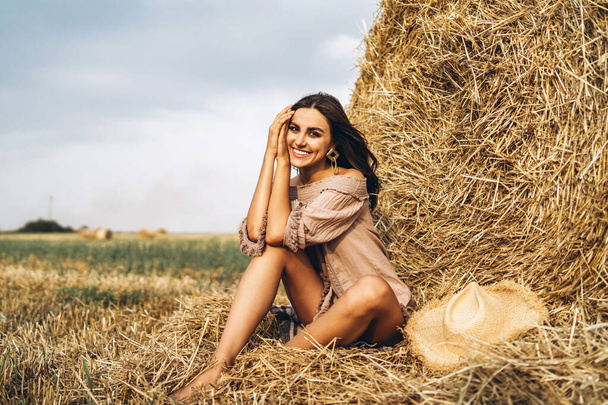 A young woman with long hair and in a dress sits near a hay bale. Woman posing smiling and looking at camera. - Fotografie, Obrázek