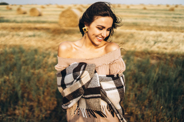 Portrait of a beautiful brunette in a dress and with a warm plaid. Woman enjoying a walk in a wheat field with hay bales. - Photo, image