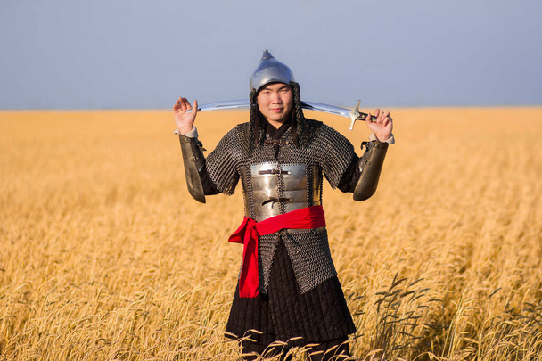 Een nomadenkrijger in oosterse harnas met een sabel en een helm uit de 16e en 18e eeuw staat tot zijn knieën in een tarweveld. - Foto, afbeelding