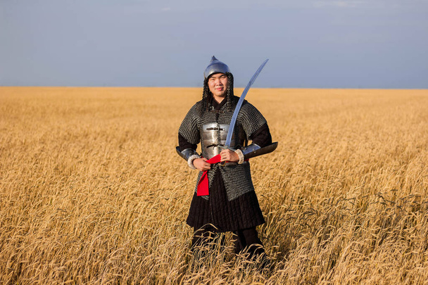 A nomad warrior in oriental armor with a saber and a helmet from the 16th and 18th centuries stands knee-deep in a wheat field. - Foto, imagen