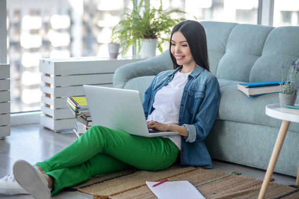 Yeşil pantolonlu genç bir kadın yerde oturuyor ve internette sohbet ediyor. - Fotoğraf, Görsel