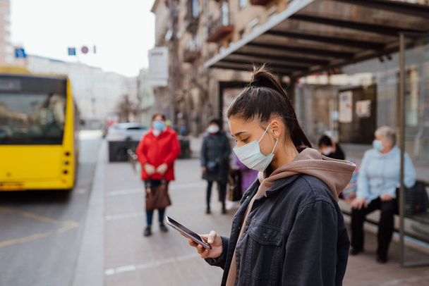 Jonge vrouw draagt chirurgische masker buiten bij bushalte in de straat - Foto, afbeelding