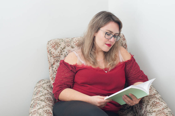 若いブロンドの女性が椅子に座って本を読んでる。彼女は赤いシャツと眼鏡をかけている。素晴らしいコピースペース. - 写真・画像