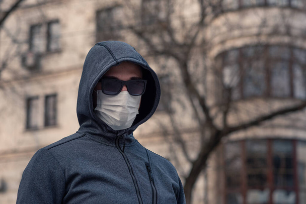portrait of a brunette man in a surgical dressing and black glasses against the backdrop of a modern building, coronavirus, disease, infection, quarantine, medical mask - 写真・画像