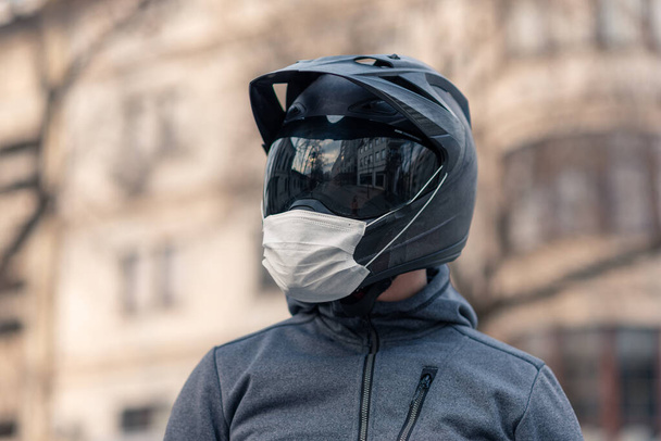 Porträt eines Mannes mit Helm. auf einem Motorradhelm eine weiße medizinische Maske. Foto auf der Straße vor dem Hintergrund von Häusern. Coronavirus, Krankheit, Infektion, Quarantäne, Covid-19 - Foto, Bild