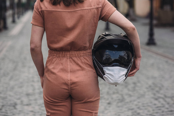 girl stands in overalls and holds in his hand a motorcycle helmet with a medical mask. Photo without a face. Against the background of the street and road. coronavirus, disease, infection, quarantine, covid-19 - Fotó, kép