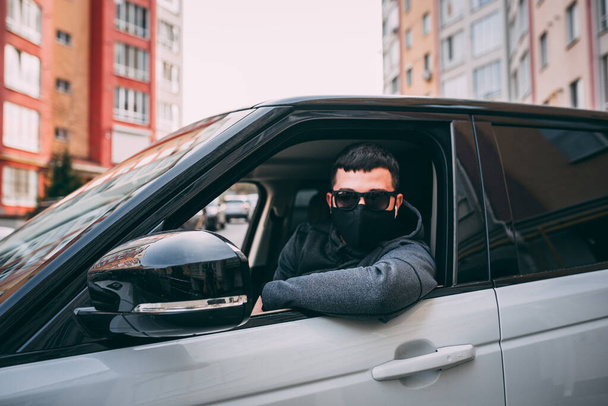 man in a black medical mask. handsome brunette driving a car, sitting at the wheel of an expensive car. thief, steal, coronavirus, disease, infection, quarantine, medical mask, covid-19 - Photo, image