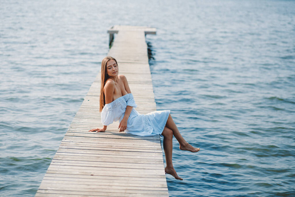 Young beautiful girl in a white summer dress went to the lake along a long wooden pier, summer the sun shines - Φωτογραφία, εικόνα
