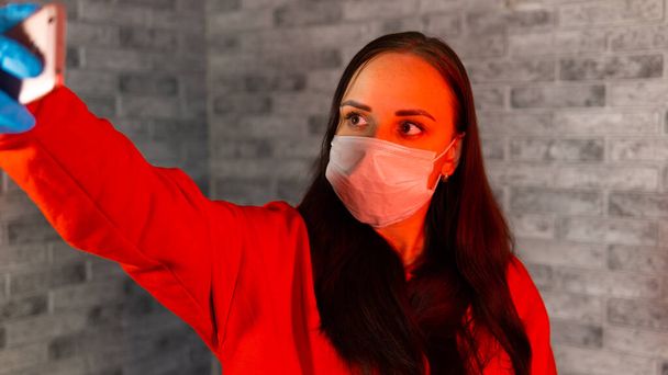 Young woman in medical mask and gloves taking selfie on smartphone, standing on gray brick background. Portrait of female spending time weekend at home and photographing of herself on mobile phone. - Фото, изображение