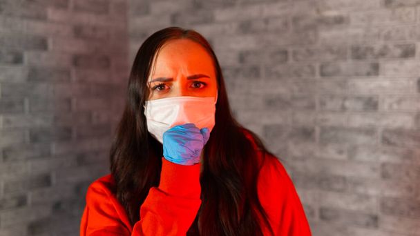 Young woman in medical mask and gloves coughing into fist, being isolated at home. Adult female protecting yourself from diseases. Concept of threat of coronavirus epidemic infection. - Фото, изображение