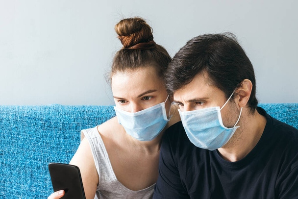 Mavi tıbbi maskeli bir çift akıllı telefondan bir şeye bakıyor. Bir erkek ve bir kadın, iki kişilik bir aile, karantina sırasında evde kalırlar. - Fotoğraf, Görsel