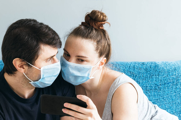 Mavi tıbbi maskeli bir çift akıllı telefona bakıyor. Erkek ve kadın, iki kişilik aile, internetten arkadaşlarını ya da ailelerini ara, selfie çek, karantina sırasında video veya haber izle - Fotoğraf, Görsel
