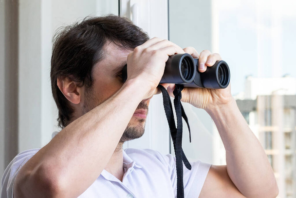 Pohledný muž hledící dalekohledem do okna s výhledem na sousední dům pro pozorování sousedů, prostředí nebo přírody během karantény. Selektivní soustředění. Detailní pohled - Fotografie, Obrázek