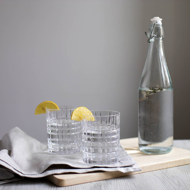 Lemon lemonade with tarragon in transparent bottle on table against background of gray wall. Two corrugated glasses with lemon slices. Gray napkin and cutting board. Copy space. Light color picture - Zdjęcie, obraz