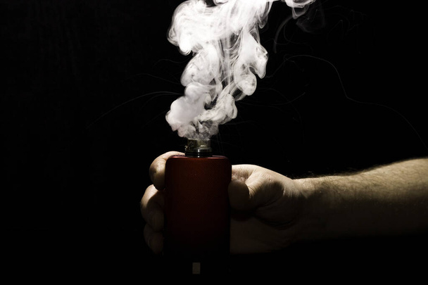 VAPE na mão de um homem com uma nuvem de vapor espesso em um fundo preto
. - Foto, Imagem