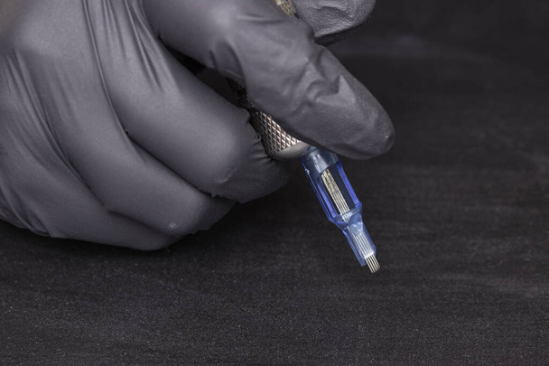 Parte de una máquina rotativa de tatuaje con una aguja en la mano en un guante negro sobre un fondo negro. Vista lateral
 - Foto, imagen